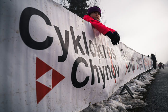 A spectator leans on the Cyklokros Chýnov banner by the course
